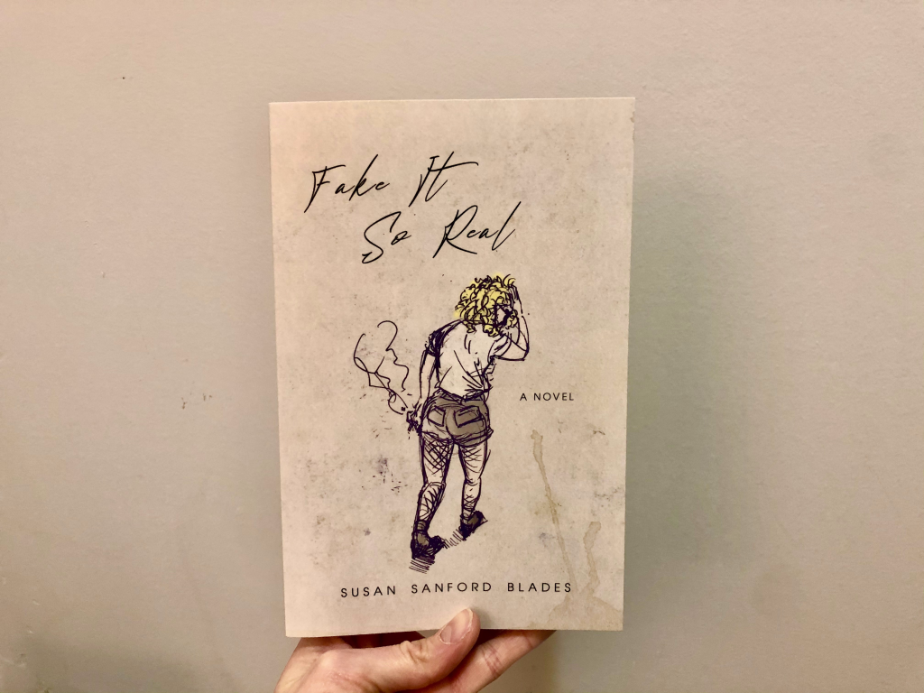 hand holding fake it so real by susan Sanford blades book cover against off-white wall